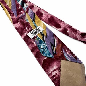 May include: A Luciano Soprani necktie featuring an abstract pattern in shades of burgundy, gold, purple, and teal. The tie has a white brand label stitched near the centre and a tan fabric lining on the back with a repeating logo pattern.