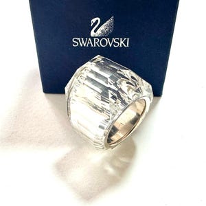 May include: A clear, faceted crystal ring with a wide band design, positioned in front of a dark blue box featuring the white Swarovski swan logo and text. The ring displays multiple geometric cut surfaces that reflect light, set against a plain background.