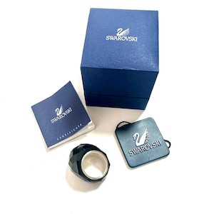 Puede incluir: Un anillo negro facetado con una banda interior plateada, acompañado de una caja azul de la marca Swarovski, una etiqueta cuadrada azul con el logotipo del cisne de Swarovski y un pequeño folleto blanco con la palabra Certificate y el logo.