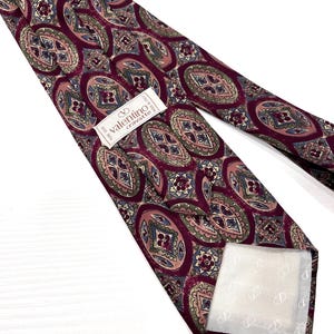 Può includere: Una cravatta bordeaux con un motivo ripetitivo di forme ovali e a rombo nei toni del verde, blu e rosa. Un'etichetta bianca è cucita sul retro con il testo "valentino cravatte" e "MADE IN ITALY". La fodera è di colore bianco.