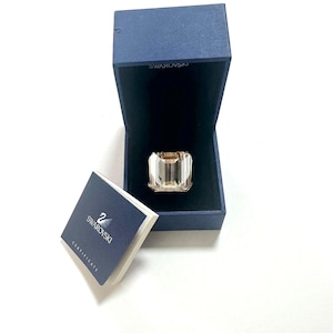 May include: A large, emerald-cut, light champagne-colored crystal ring sits inside a dark blue, velvet-lined Swarovski box. Beside the box is a small, dark blue folded booklet with the Swarovski swan logo and the word CERTIFICATE printed on it.
