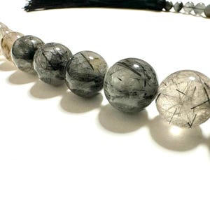 Könnte beinhalten: Eine Kette aus runden, polierten Rutilquarzperlen mit sichtbaren schwarzen, nadelartigen Einschlüssen im klaren und grauen Stein. Die Perlen sind in einer geschwungenen Linie angeordnet, die von dunkleren Grautönen zu helleren, durchscheinenden Nuancen übergeht, und mit einer schwarzen Quaste versehen.