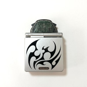 Puede incluir: Una consola de juegos portátil plateada con un diseño gráfico tribal negro en la cubierta superior y el logotipo de Nintendo impreso en la parte inferior. Un cartucho de juego gris oscuro translúcido está insertado en la ranura superior.