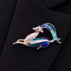 May include: A rose gold-toned brooch shaped like a leaping gazelle or antelope. The animal features textured metal, clear crystal accents along the body, and vibrant blue and teal enamel detailing on the curved horns and hindquarters.
