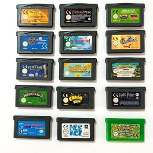 Op de afbeelding: Een verzameling van veertien Game Boy Advance-videogamecartridges gerangschikt in vijf rijen. Dertien cartridges zijn donkergrijs met rechthoekige labels met verschillende speltitels en illustraties, terwijl één cartridge felgroen is.