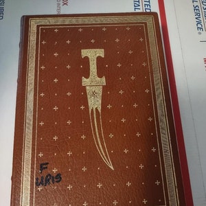 May include: A brown leather-bound book with a gold-colored design on the front cover. The design features a stylized dagger with a gold-colored handle and a silver-colored blade. The book has a gold-colored ribbon bookmark attached to the spine. The book is titled "I" in gold lettering.