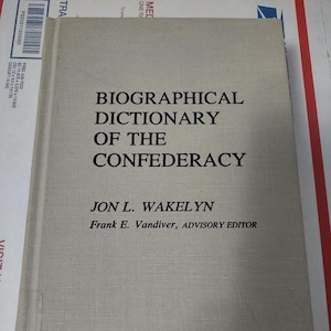 May include: A hardcover book titled "BIOGRAPHICAL DICTIONARY OF THE CONFEDERACY" by Jon L. Wakelyn. The book has a light gray cover with black text. The advisory editor is Frank E. Vandiver. The book is on a white and red surface.