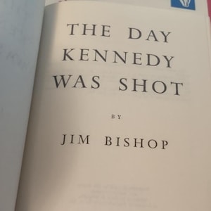 May include: Open book page with the title "THE DAY KENNEDY WAS SHOT" by Jim Bishop. Published by Funk & Wagnalls, New York. The text is in a classic serif font, printed in black on a cream-colored paper.