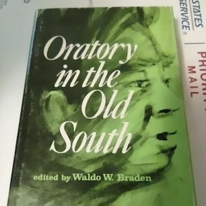 May include: A green book cover with a portrait of a person on the front. The title of the book is "Oratory in the Old South" and the author is Waldo W. Braden.