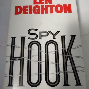 May include: A white book cover with the title "SPY HOOK" in black and gray letters with barbed wire behind the letters. The author's name, "LEN DEIGHTON", is printed in red letters above the title.
