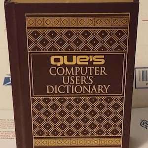 May include: A hardcover book titled "Que's Computer User's Dictionary." The book has a dark brown cover with gold geometric patterns and text. The title is in gold lettering. The book is a reference guide for computer users.