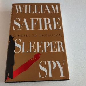 May include: A gold book cover with the title "Sleeper Spy" by William Safire. The book cover features a silhouette of a person in a trench coat walking away from a red shadow.