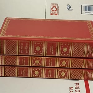 May include: Three red hardcover books with gold accents and titles. The top book is titled "The Prince & The Discourses" by Niccolo Machiavelli. The middle book is titled "Autobiography" by Benjamin Franklin. The bottom book is titled "Poor Richard's Almanack" by Benjamin Franklin.
