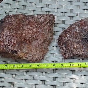 May include: Two rough, reddish-brown rocks of varying sizes are displayed on a light gray woven surface. A yellow measuring tape is placed next to the rocks, showing measurements in inches. The larger rock is approximately 6 inches long.