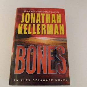 May include: A red, white, and blue book cover with the title "Bones" in large, bold, white letters. The author's name, "Jonathan Kellerman", is printed in yellow letters above the title. The book is described as "An Alex Delaware Novel".