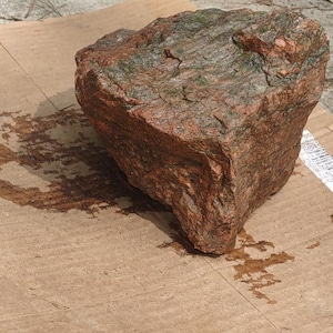 May include: A large, rough-textured rock with reddish-brown and green hues. The rock is irregularly shaped and sits on a piece of cardboard. The surface of the cardboard has water stains.