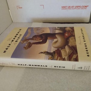May include: The Half-Mammals of Dixie book cover features a colorful illustration of a mermaid-like creature with a human torso and a fish tail. The creature is sitting on a pile of books and holding a small object in its hand. The book title is written in large, bold letters on the cover.