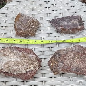 May include: Four irregularly shaped rocks in shades of brown, gray, and red are displayed on a white woven surface. A yellow measuring tape is placed across the rocks, showing measurements up to 14 inches. The rocks vary in size and texture.