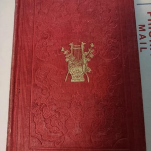 May include: A red hardcover book with a gold embossed design on the front cover. The design features a lyre, a musical instrument, surrounded by floral motifs.