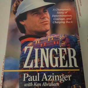 May include: A book titled "Zinger" by Paul Azinger with Ken Abraham. The cover features a color photo of Paul Azinger wearing a white visor and a colorful shirt. The book title is in large, bold, white letters. The subtitle is "A Champion's Story of Determination, Courage, and Charging Back".