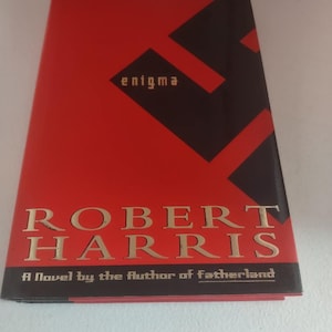 May include: A book cover for "Enigma" by Robert Harris. The cover is predominantly red with a large black geometric design. The title and author's name are in gold lettering. The text "A Novel by the Author of Fatherland" is also visible.