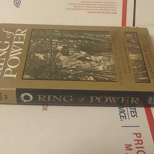 K&ouml;nnte beinhalten: Ein gebundenes Buch mit dem Titel Ring of Power von Jean Shinoda Bolen, M.D. Der Einband hat einen goldenen Hintergrund mit einer zentralen schwarz-wei&szlig;en Illustration einer Figur im Wald. Der schwarze Buchr&uuml;cken zeigt Titel und Autor in Wei&szlig;.