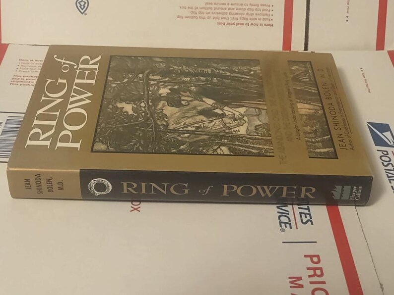 K&ouml;nnte beinhalten: Ein gebundenes Buch mit dem Titel Ring of Power von Jean Shinoda Bolen, M.D. Der Einband hat einen goldenen Hintergrund mit einer zentralen schwarz-wei&szlig;en Illustration einer Figur im Wald. Der schwarze Buchr&uuml;cken zeigt Titel und Autor in Wei&szlig;.