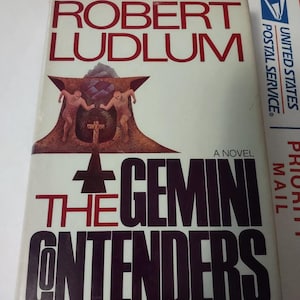 May include: A white book cover with black text that reads "Robert Ludlum" and "The Gemini Contenders." The book cover features a black and white illustration of two figures standing on a platform.