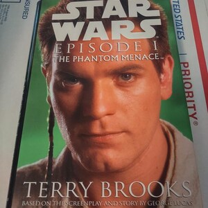May include: A book cover featuring a close-up of a man's face with a green background. The title is "Star Wars Episode I The Phantom Menace" and the author is "Terry Brooks". The book is based on the screenplay and story by George Lucas.