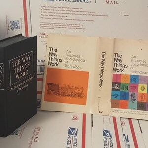 May include: Two black hardcover books titled "The Way Things Work" with an illustrated encyclopedia of technology. The books are on a white surface with a stack of brown cardboard boxes behind them.