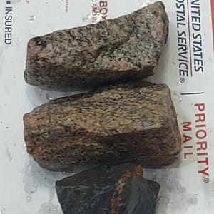 May include: Three rough-hewn rocks with varying textures and colors. Two are primarily brown and gray, while the third is dark gray with a reddish-orange streak. The rocks are on a white surface with USPS Priority Mail branding.
