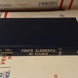 May include: A navy blue hardcover book titled "Finite Elements in Fluids, Vol. 2" by Wiley. The book's spine displays the title and author's names. The book is resting on a white and red USPS Priority Mail box.