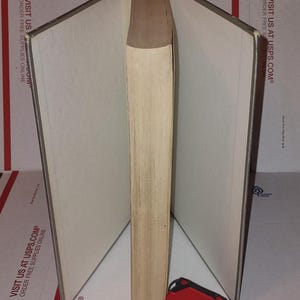 Puede incluir: Un libro abierto con p&aacute;ginas blanquecinas y un lomo marr&oacute;n claro. Una cinta m&eacute;trica roja y negra descansa en la base del libro. El libro est&aacute; sobre una superficie con texto impreso.