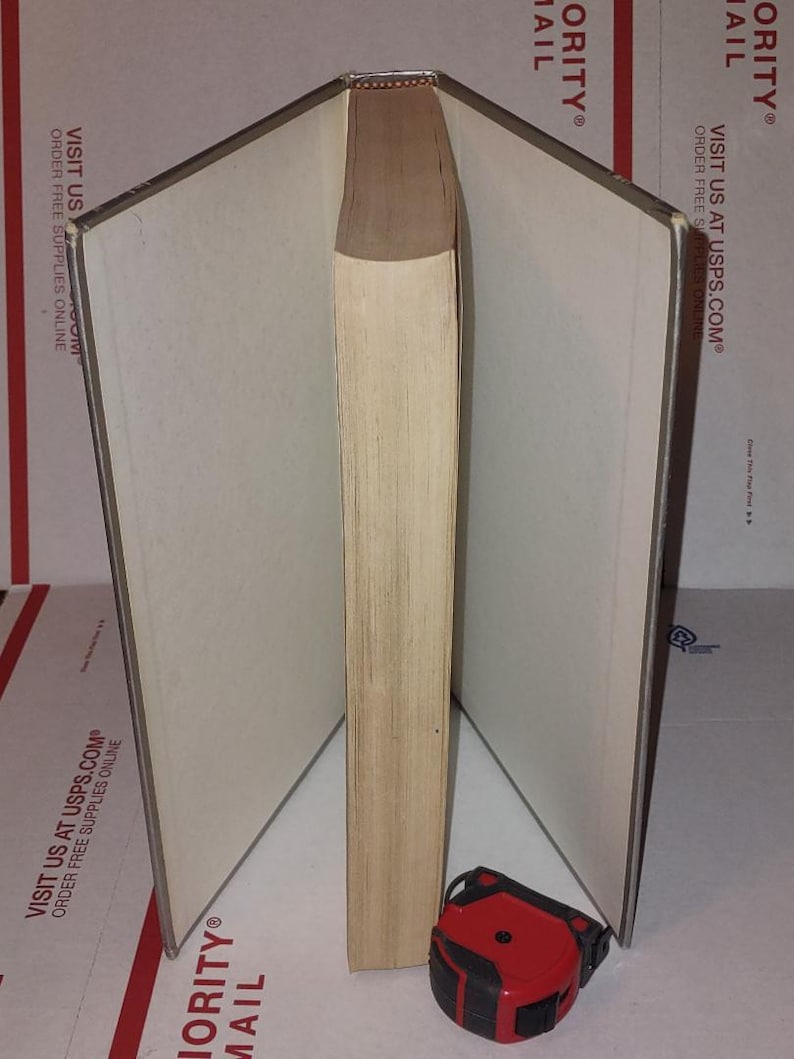 Puede incluir: Un libro abierto con p&aacute;ginas blanquecinas y un lomo marr&oacute;n claro. Una cinta m&eacute;trica roja y negra descansa en la base del libro. El libro est&aacute; sobre una superficie con texto impreso.