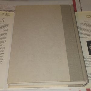K&ouml;nnte beinhalten: Ein gebundenes Buch mit hellbeigem Vorderdeckel und einem Buchr&uuml;cken aus strukturiertem grauem Stoff. Das Buch liegt flach und zeigt den Vorderdeckel sowie den Buchr&uuml;cken, w&auml;hrend die R&uuml;ckseite nicht sichtbar ist. Schlichtes, einfarbiges Design.