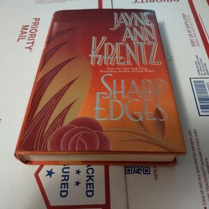 May include: A paperback book titled "Sharp Edges" by Jayne Ann Krentz. The cover features a red rose with a background of orange and yellow flames.