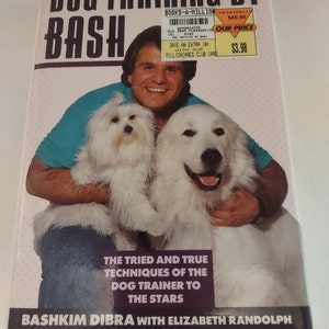 May include: A book cover titled "DOG TRAINING BY BASH" featuring a man holding two white dogs. The cover includes the text "THE TRIED AND TRUE TECHNIQUES OF THE DOG TRAINER TO THE STARS" and the author's name, Bashkim Dibra.