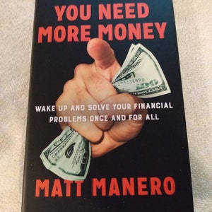 May include: Book cover with the title "YOU NEED MORE MONEY" in red letters on a black background. A hand is squeezing several one hundred dollar bills. The author's name, Matt Manero, is also in red.