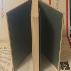 K&ouml;nnte beinhalten: Ein Hardcover-Buch, das aufrecht steht, wobei die Buchdeckel nach au&szlig;en ge&ouml;ffnet sind und dunkelgr&uuml;ne Vorsatzbl&auml;tter zeigen. Der Buchr&uuml;cken ist nach vorne gerichtet und zeigt die cremefarbenen Seiten des Buchblocks. Das Buch steht vor einem wei&szlig;en Hintergrund.