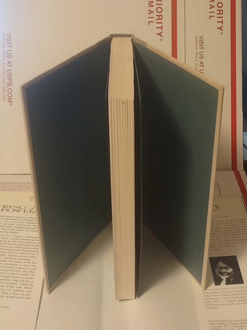 K&ouml;nnte beinhalten: Ein Hardcover-Buch, das aufrecht steht, wobei die Buchdeckel nach au&szlig;en ge&ouml;ffnet sind und dunkelgr&uuml;ne Vorsatzbl&auml;tter zeigen. Der Buchr&uuml;cken ist nach vorne gerichtet und zeigt die cremefarbenen Seiten des Buchblocks. Das Buch steht vor einem wei&szlig;en Hintergrund.