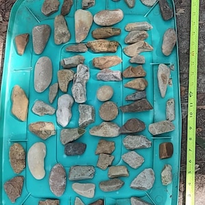 May include: A collection of various-sized rocks and stones in shades of gray, brown, and beige, arranged on a turquoise plastic tray. A yellow measuring tape is visible along the right side, showing measurements in inches.