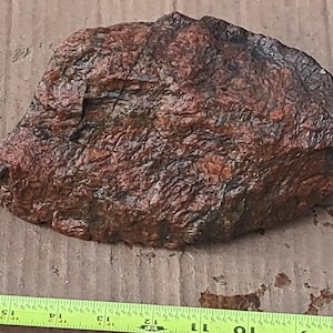 May include: A rough, reddish-brown rock with a textured surface. The rock has dark brown and black areas, and is irregularly shaped. A yellow and green measuring tape is at the bottom.