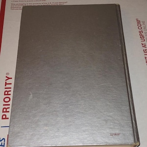 Puede incluir: Un libro plateado con una cubierta texturizada. El libro es rectangular y tiene un lomo. El n&uacute;mero "325697" est&aacute; impreso en la esquina inferior derecha. El libro est&aacute; sobre una superficie a rayas blancas y rojas.