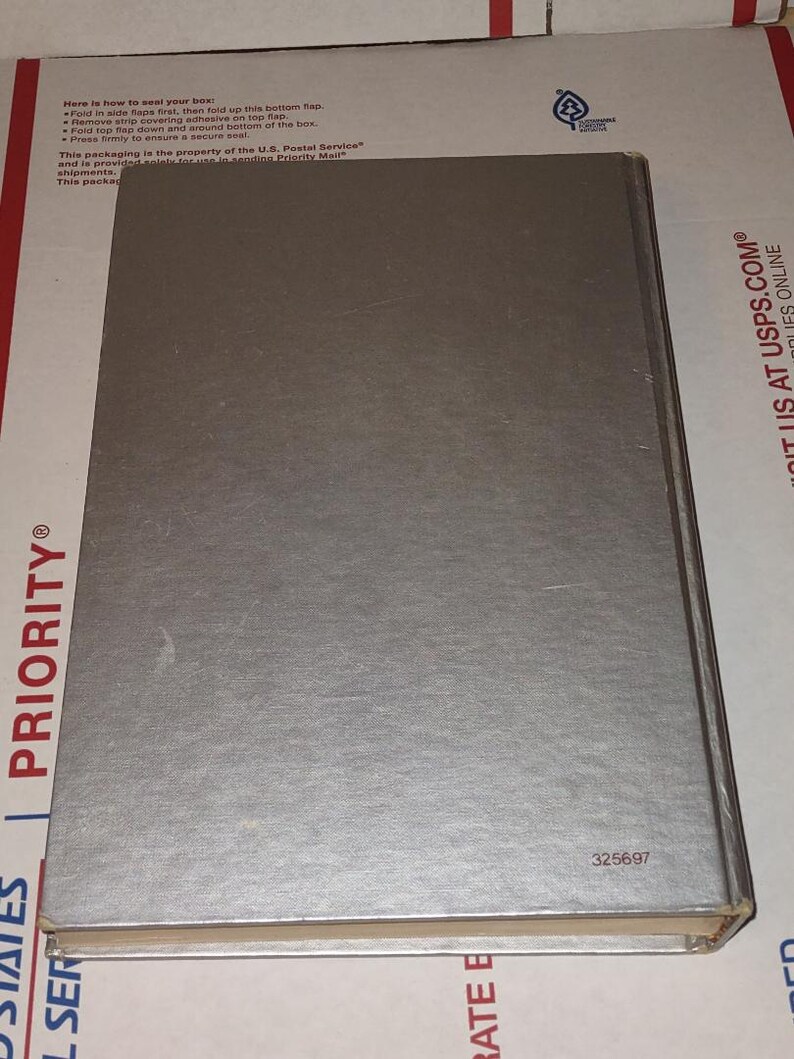 Puede incluir: Un libro plateado con una cubierta texturizada. El libro es rectangular y tiene un lomo. El n&uacute;mero "325697" est&aacute; impreso en la esquina inferior derecha. El libro est&aacute; sobre una superficie a rayas blancas y rojas.