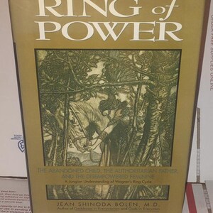K&ouml;nnte beinhalten: Ein gebundenes Buch mit dem Titel Ring of Power von Jean Shinoda Bolen, M.D. Der Einband hat einen goldenen Hintergrund mit wei&szlig;em Text und einer zentralen Illustration einer Person mit einem Pferd in einem Wald. Der Untertitel lautet The Abandoned Child, The Authoritarian Father, And The Disempowered Feminine, A Jungian Understanding of Wagner's Ring Cycle.