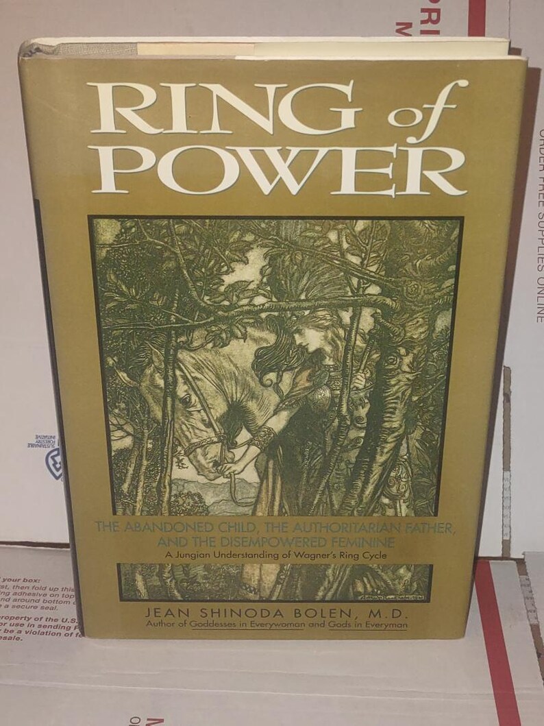 K&ouml;nnte beinhalten: Ein gebundenes Buch mit dem Titel Ring of Power von Jean Shinoda Bolen, M.D. Der Einband hat einen goldenen Hintergrund mit wei&szlig;em Text und einer zentralen Illustration einer Person mit einem Pferd in einem Wald. Der Untertitel lautet The Abandoned Child, The Authoritarian Father, And The Disempowered Feminine, A Jungian Understanding of Wagner's Ring Cycle.