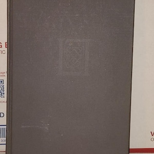May include: A hardcover book with a gray cover. The cover has a subtle embossed design in the center. The book is rectangular and appears to be in good condition, with minimal wear visible. The book is placed on a white and red surface.