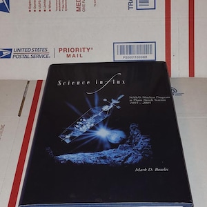 May include: A hardcover book titled "Science in Flux" with a dark blue cover. The book features an illustration of a spacecraft and a celestial body. The author's name, Mark D. Bowles, is printed on the cover.
