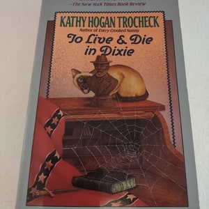 May include: Book cover with a blue background and a title that reads "To Live & Die in Dixie." The cover features a cat sitting on a table with a man in a Confederate uniform. The text "A Callahan Garrity Mystery" is at the bottom of the cover.