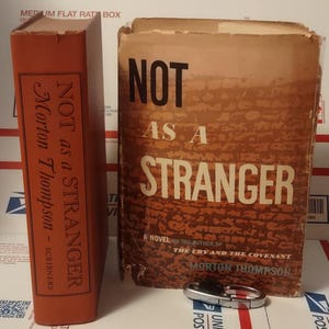 May include: Two vintage books, "Not as a Stranger" by Morton Thompson, are displayed. The book on the left is orange with white text. The other book has a brown cover with the title in large black letters. A small silver object rests in front.